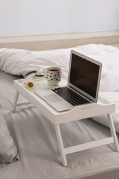 White tray table with laptop, cup of drink and daisy on bed indoors