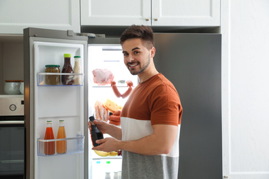 Young man with bottle of sauce near open refrigerator indoors