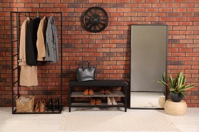 Photo of Stylish hallway with coat rack, mirror and shoe storage bench near brick wall. Interior design