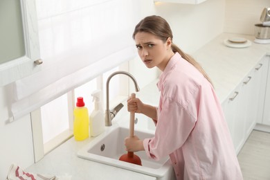 Upset young woman using plunger to unclog sink drain in kitchen