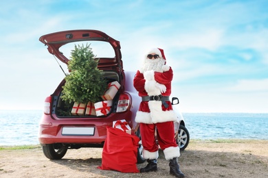Authentic Santa Claus near red car with gift boxes and Christmas tree on beach