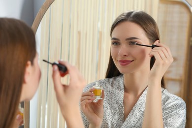 Photo of Young woman applying oil on her eyelashes near mirror indoors