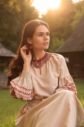 Photo of Beautiful woman wearing embroidered dress in village on sunny day. Ukrainian national clothes