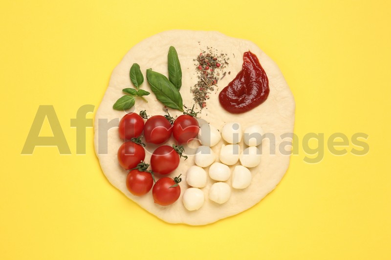Photo of Raw dough and other ingredients for pizza on yellow background, top view