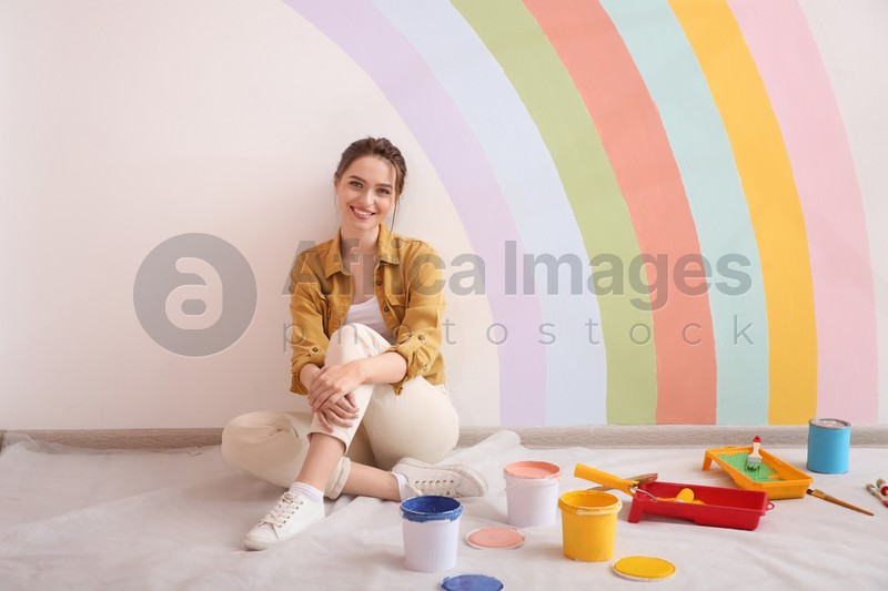 Young woman and decorator's tools near wall with painted rainbow indoors