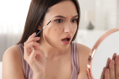 Beautiful woman with fallen eyelashes and cosmetic brush looking into mirror indoors