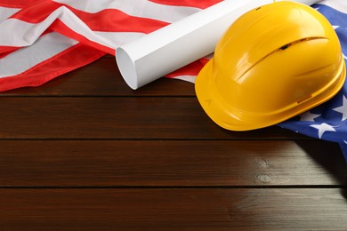 Photo of Yellow protective hard hat, construction drawing and American flag on wooden table. Space for text