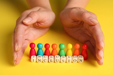 Woman protecting colorful pawns and wooden cubes with word Inclusion on yellow background, closeup