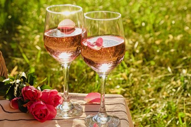 Photo of Glasses of delicious rose wine with petals and flowers on white picnic blanket outside