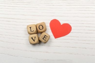Mini cubes with letters forming word Love near red paper heart on white wooden background, flat lay