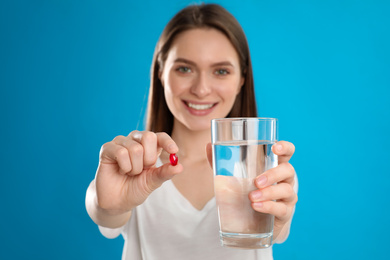 Photo of Young woman with vitamin pill and glass of water against blue background, focus on hands