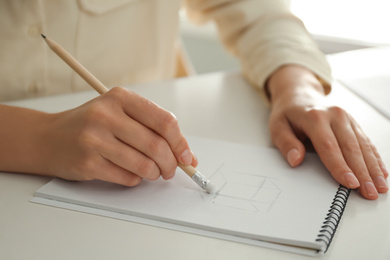 Woman correcting picture in notepad with pencil eraser at white table, closeup