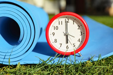 Photo of Alarm clock and fitness mat on green grass outdoors, closeup. Morning exercise