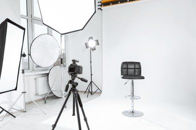 Photo of Tripod with camera, bar stool and professional lighting equipment in modern photo studio