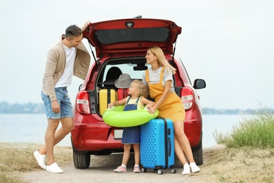 Happy family with suitcases and inflatable ring near car on riverside