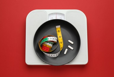 Scales with plate, weight loss pills and measuring tape on red background, top view