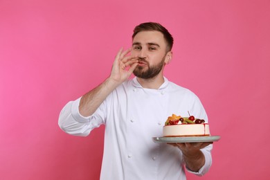 Photo of Happy professional confectioner in uniform with cake showing delicious gesture on pink background