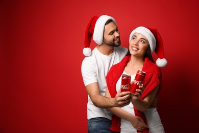 MYKOLAIV, UKRAINE - JANUARY 27, 2021: Young couple in Christmas hats holding cans of Coca-Cola on red background. Space for text