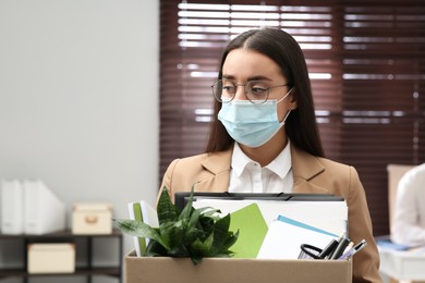 Upset dismissed woman wearing protective mask carrying box with personal stuff in office