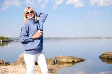 Happy woman in stylish sweater with cup of coffee on beach