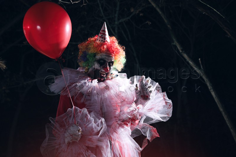 Photo of Terrifying clown with red air balloon outdoors at night. Halloween party costume