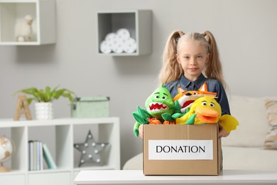 Cute little girl holding donation box with soft toys at home, space for text