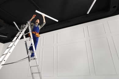 Electrician in uniform installing ceiling lamp indoors, low angle view