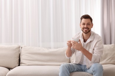 Photo of Happy man drinking coffee while resting on sofa near window with beautiful curtains in living room. Space for text