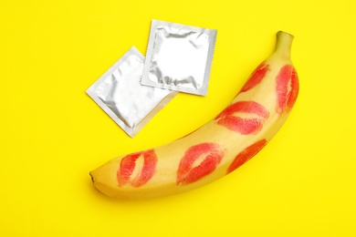 Condoms and banana with lipstick kiss marks on yellow background, flat lay. Safe sex