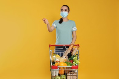 Woman with protective mask and shopping cart full of groceries on yellow background