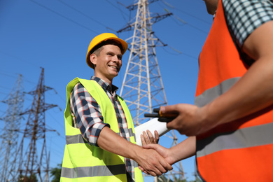 Professional electricians shaking hands near high voltage tower, closeup
