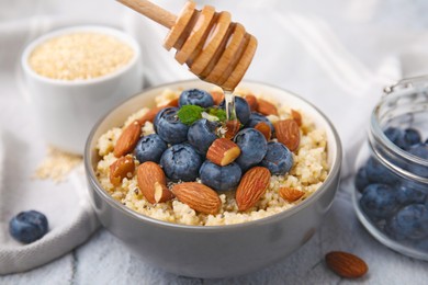 Pouring honey onto delicious cooked quinoa with almonds and blueberries in bowl on white textured table, closeup