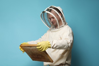 Photo of Beekeeper in uniform holding hive frame with honeycomb on light blue background