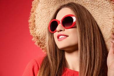 Young woman wearing stylish sunglasses and hat on red background