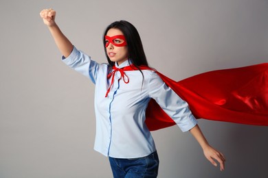 Confident young woman wearing superhero cape and mask on light grey background