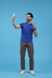 Photo of Smiling man taking selfie with smartphone and showing peace sign on light blue background