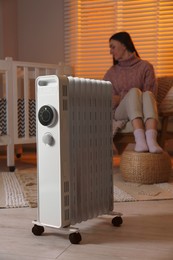 Photo of Young woman near crib and modern portable electric heater indoors, focus on device