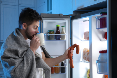 Young man taking sausages out of refrigerator at night