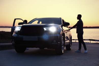 Young man standing near family car at sunset
