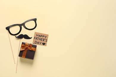 Photo of Card with phrase Happy Father's Day, paper glasses, mustache and gift box on pale yellow background, flat lay. Space for text