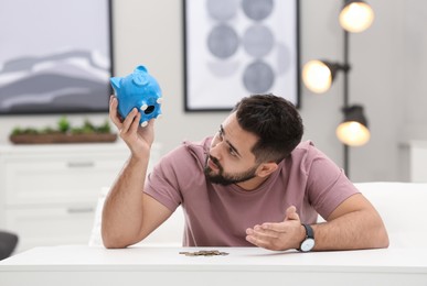 Worried young man with piggy bank and money at white table indoors