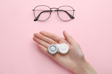 Woman holding case with contact lenses near glasses on pink background, top view