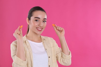 Young woman inserting foam ear plugs on pink background. Space for text