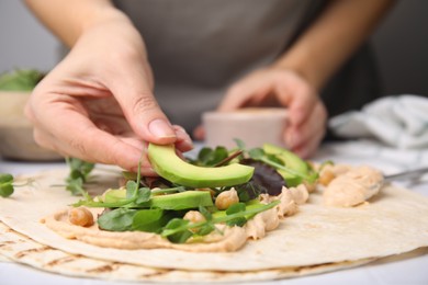 Photo of Woman making delicious hummus wrap with vegetables at table, closeup