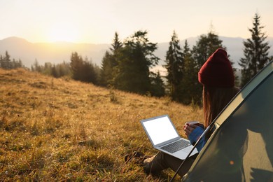 Woman working on laptop near camping tent outdoors surrounded by beautiful nature