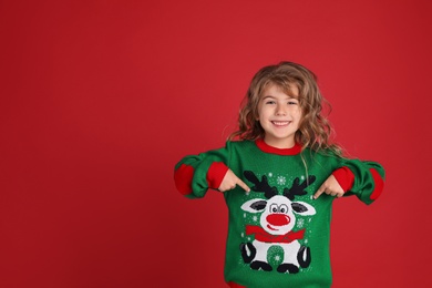 Cute little girl pointing at her green Christmas sweater against red background. Space for text