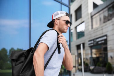Handsome young man with stylish sunglasses and backpack near building outdoors