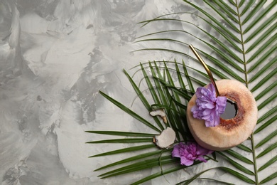 Young peeled coconut with straw, tropical leaf and flowers on grey table, flat lay. Space for text