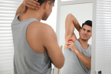 Handsome man applying deodorant to armpit near mirror at home