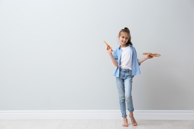 Little girl with brush and paint palette near light wall indoors. Space for text